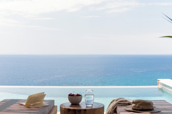 A serene infinity pool with a view of the ocean, a wooden stool with a bowl of fruit and a glass of water, and a book on a lounge chair. - Villa Lefko a Cycladic escape in the heart of the Aegean (Μέγας Γιαλός)