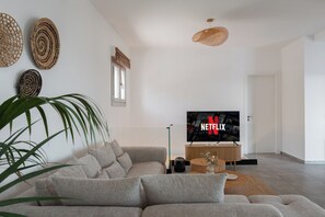 A modern living room with a grey sofa, a TV displaying the Netflix logo, and a large plant. - Villa Lefko a Cycladic escape in the heart of the Aegean (Μέγας Γιαλός)