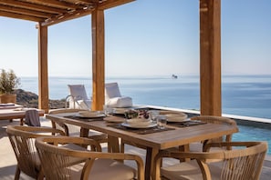 Mesa de madeira com cadeiras, vista para o mar e um navio ao longe.