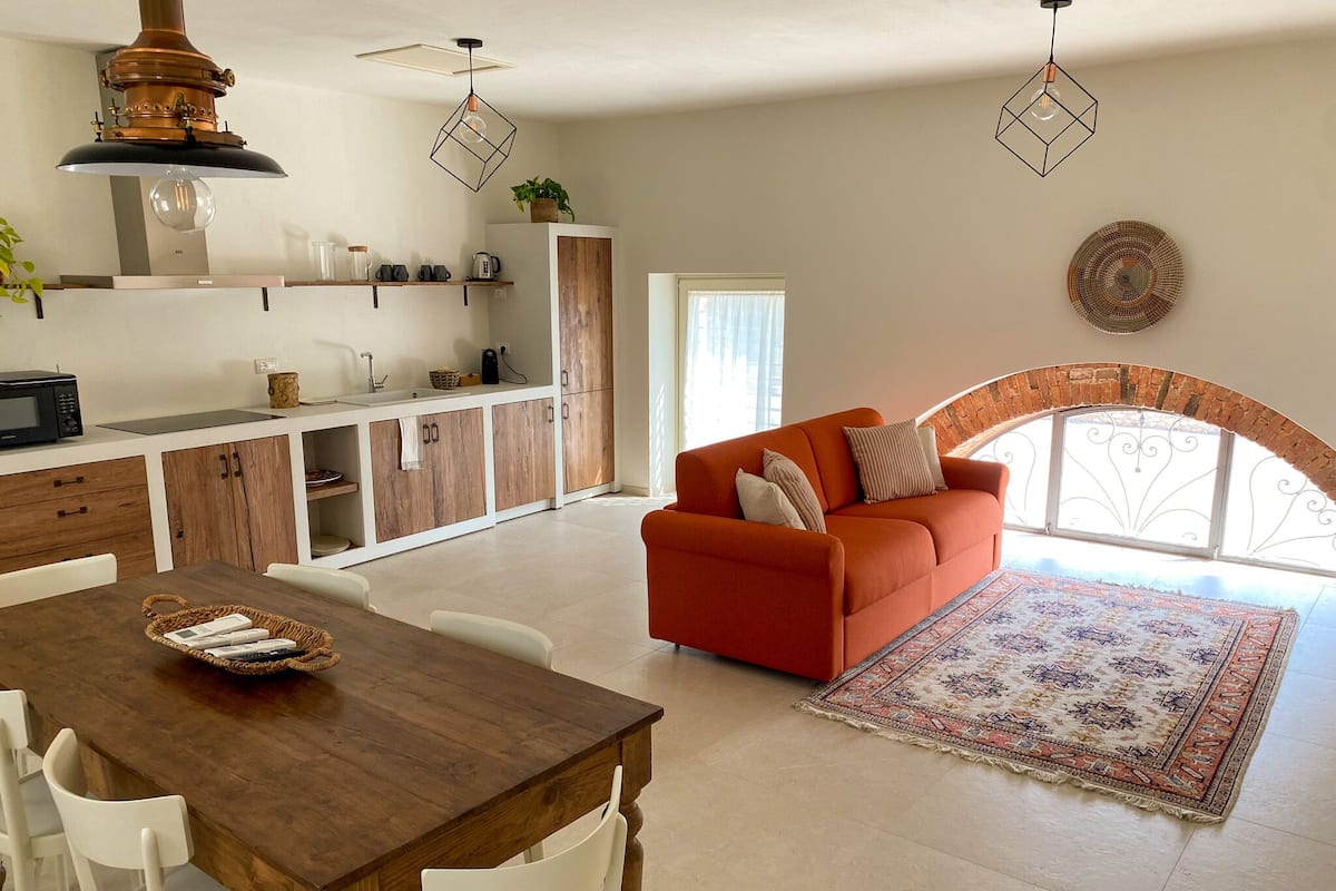 A modern kitchen with wooden cabinets, a dining table with chairs, and a sofa in a bright living area with a large window.