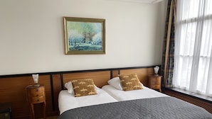 A neatly made bed with a quilted grey bedspread, two pillows with a leaf pattern, a wooden headboard, a bedside table with a lamp, and a painting of a park scene on the wall. - Logement Garjen (Harlingen)