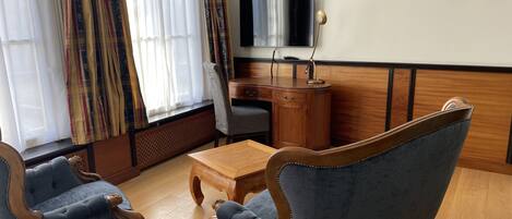 A room with two blue upholstered chairs, a wooden coffee table, a television on the wall, and a desk with a lamp.