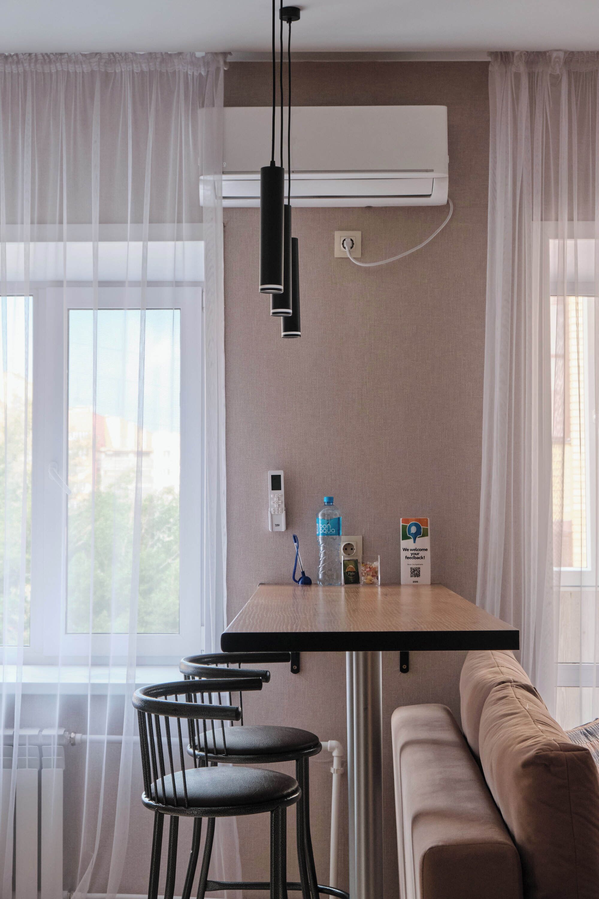 A modern room with a wooden table, two black bar stools, a beige sofa, and a large window with sheer curtains.