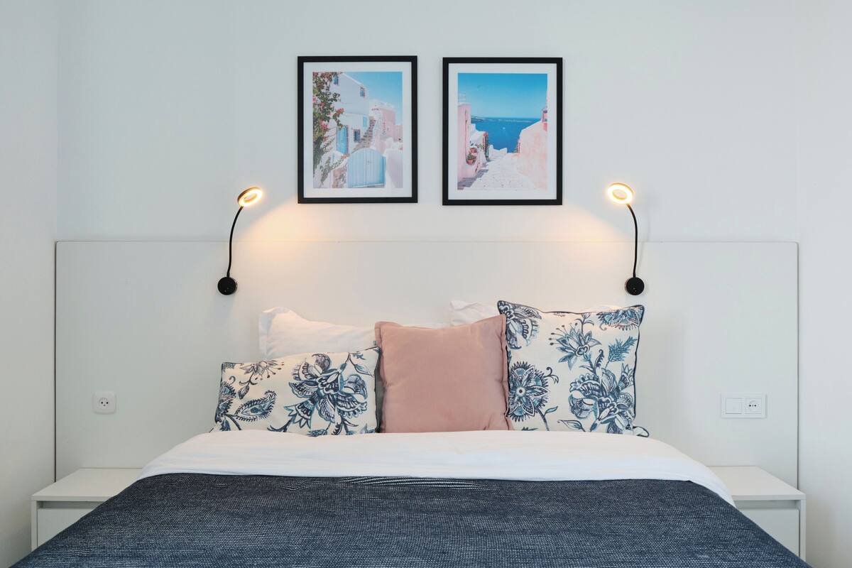 Un llit ben fet amb un coixí estampat en blau i blanc, un coixí rosa i un llit cobert amb un estovat de blau fosc. Dues fotografies emmarcades hi ha sobre el llit, una que mostra una escena costanera i l'altra un cel blau.