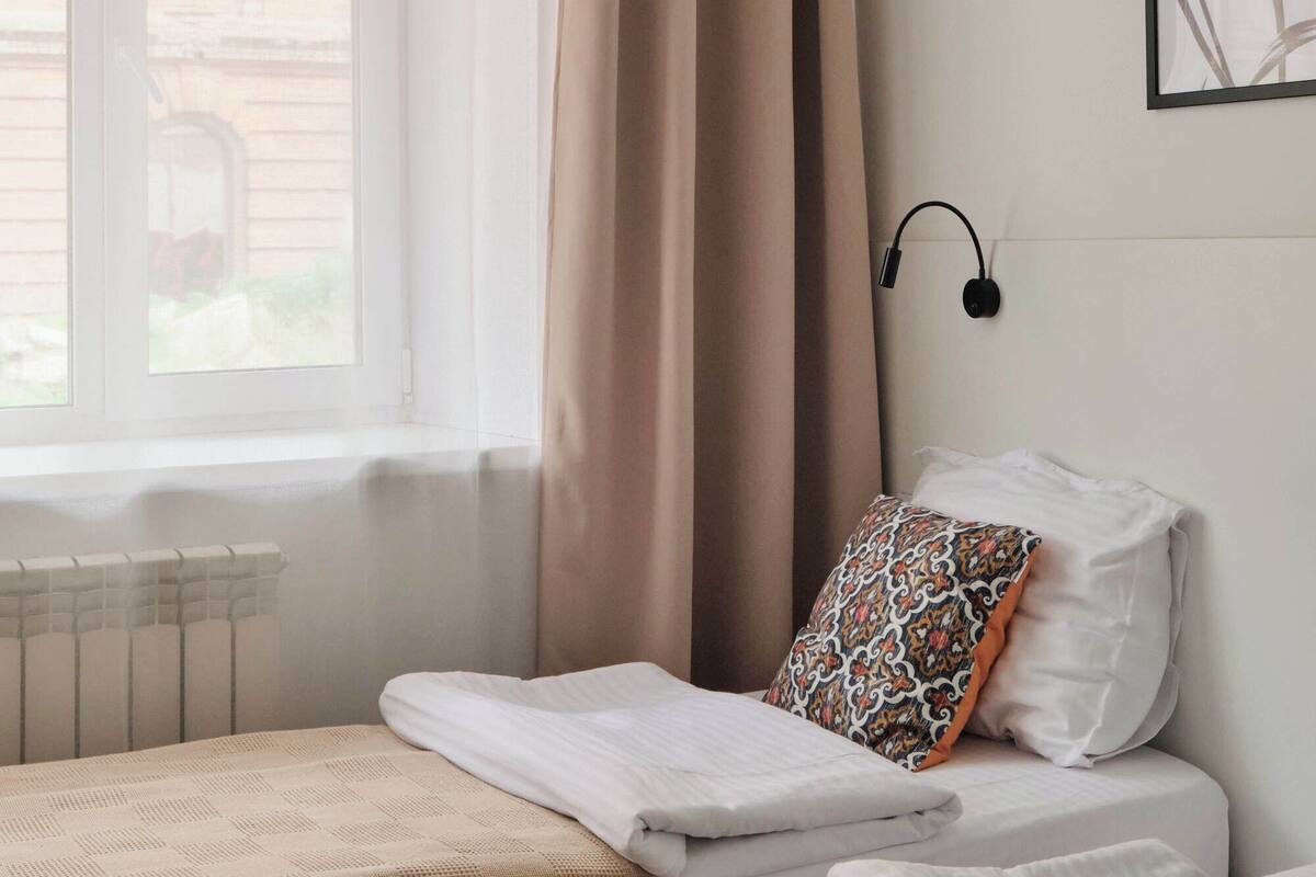 Una habitació d'hotel amb dos llits, una finestra amb cortines, un aire condicionat, una làmpada penjada a la paret i un quadre amb marc a la paret.
