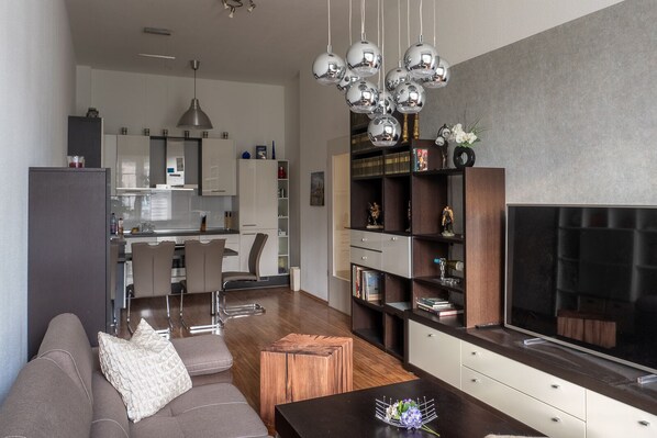 A modern living room with a sofa, coffee table, and a TV. There is a kitchen in the background with white cabinets and a dining area. - 3 room apartment "Karl May", Radebeul-Ost, 76sqm (Radebeul)