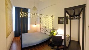 A small, cozy room with a single bed, a desk, and a chair. There is a ceiling fan, string lights, and a picture on the wall.