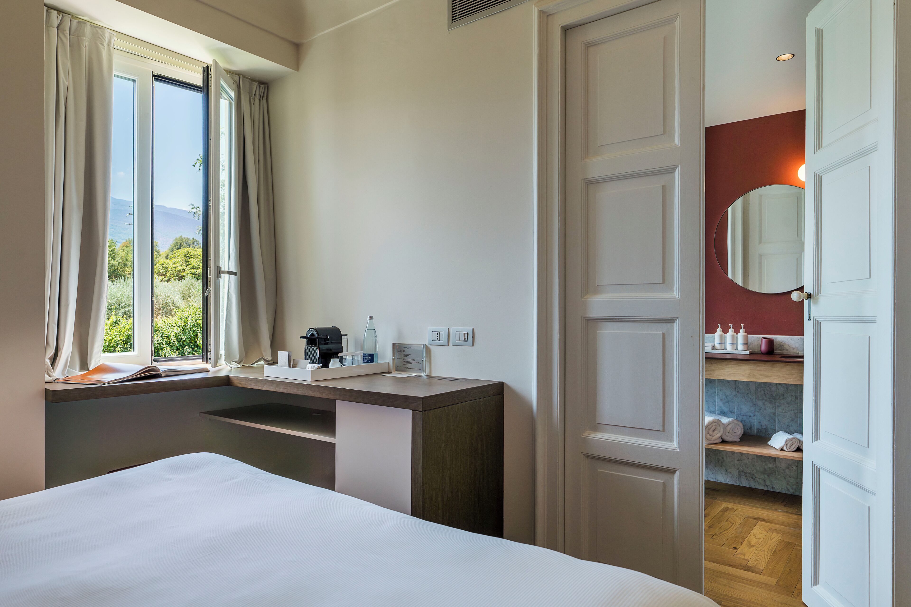 Una habitación de hotel con cama, escritorio con espejo, ventana con cortinas y vistas al exterior.