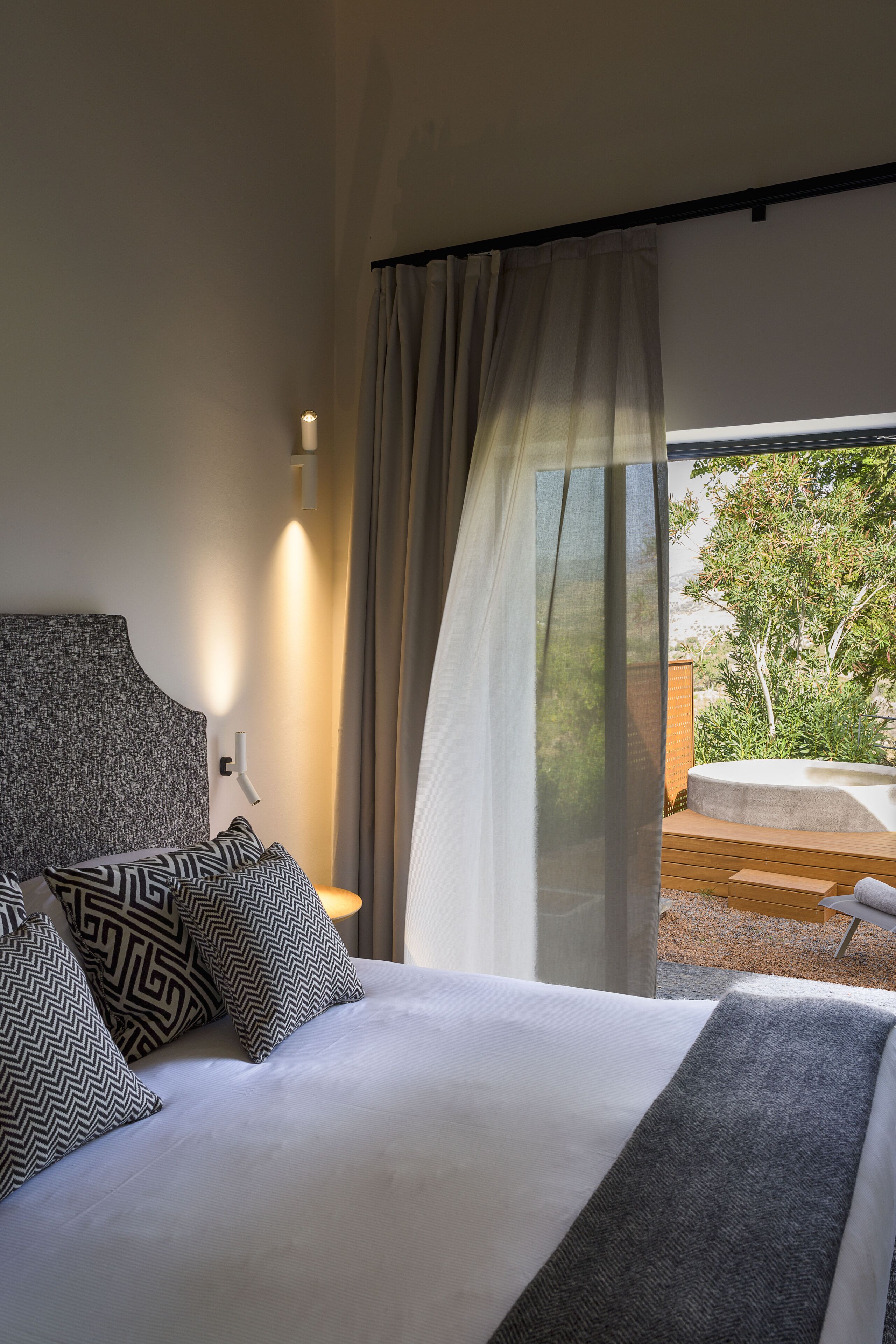 Un dormitorio moderno con una cama grande, almohadas estampadas, un cabecero y vistas a un espacio exterior a través de cortinas translúcidas.