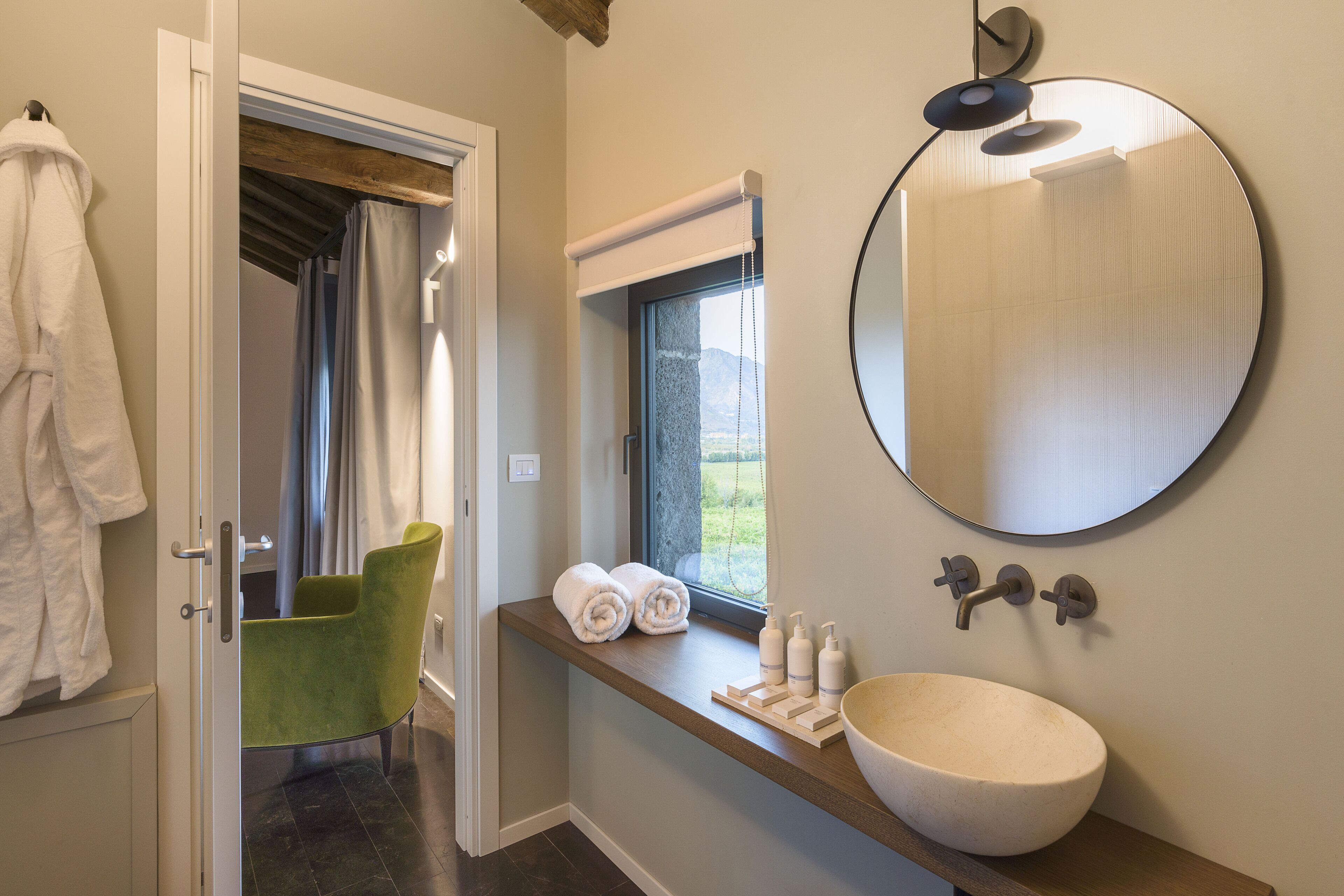 Une salle de bain avec un grand miroir rond, un meuble-lavabo en bois, une chaise verte et une vue sur l’extérieur par une fenêtre.
