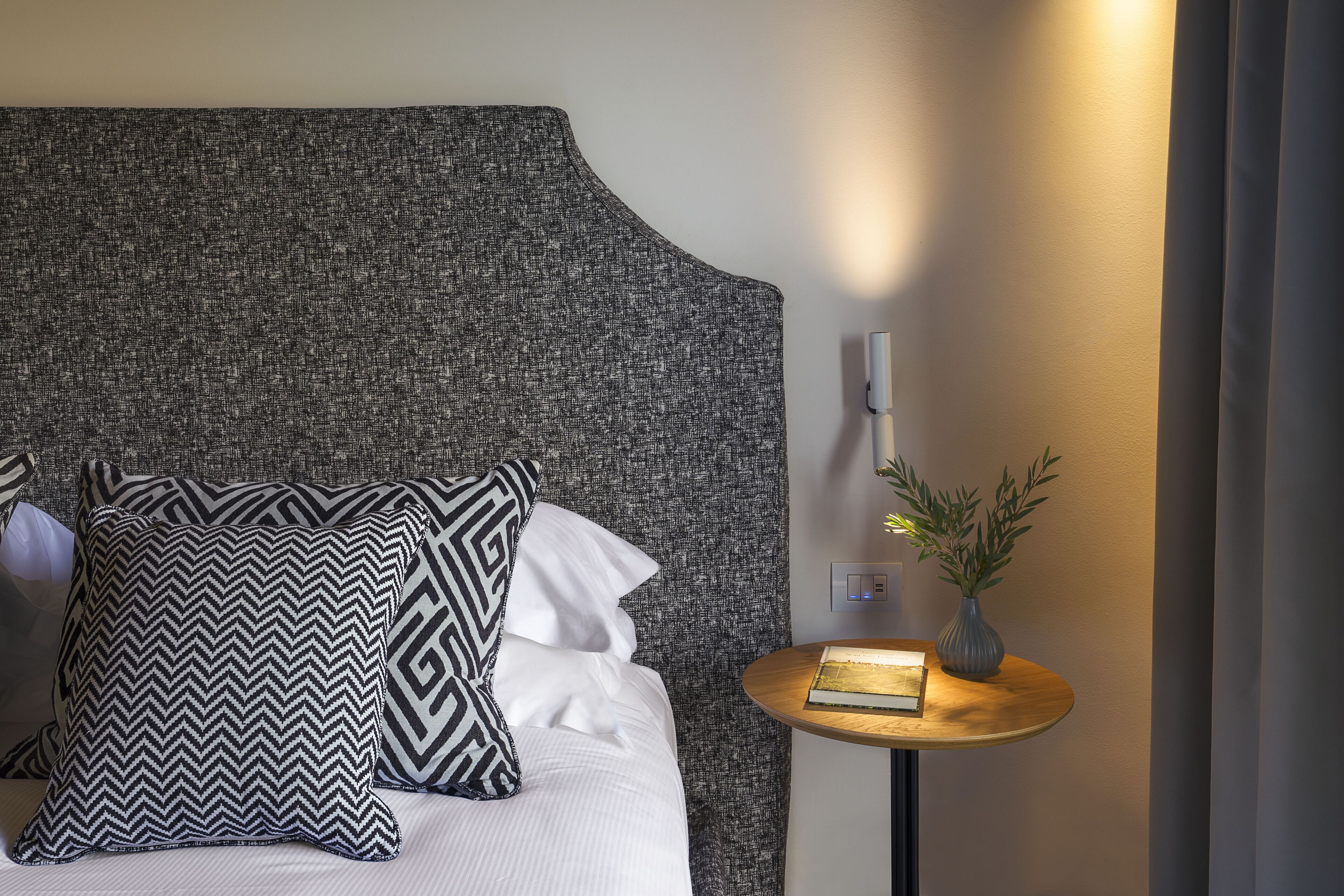 Un lit bien fait, agrémenté d’oreillers à motifs, une table de chevet avec un livre et un vase, et une lampe fixée au mur.