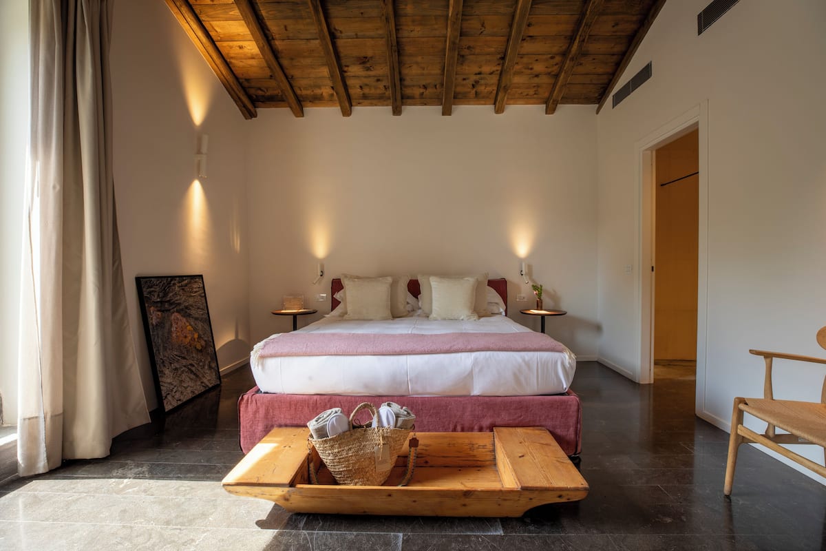 Een slaapkamer met een bed, nachtkastjes, een houten bankje, een mand en een deur.