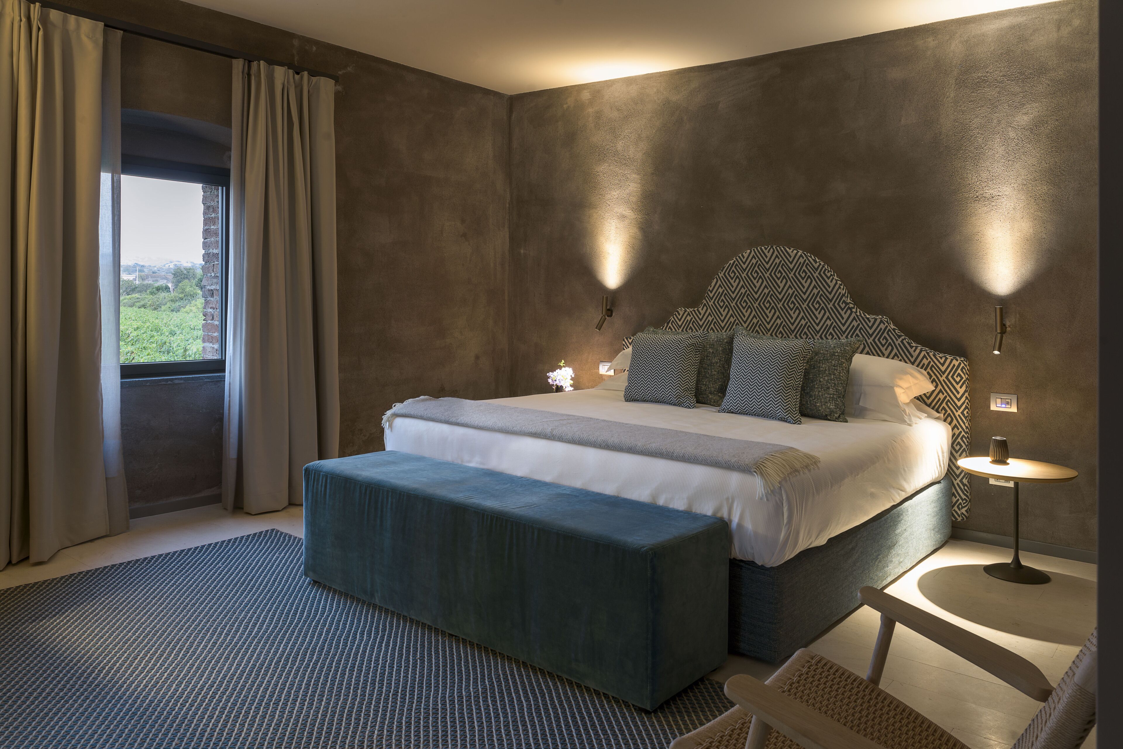 Une chambre à coucher avec un grand lit, un banc, une petite table et une vue sur la verdure par la fenêtre.