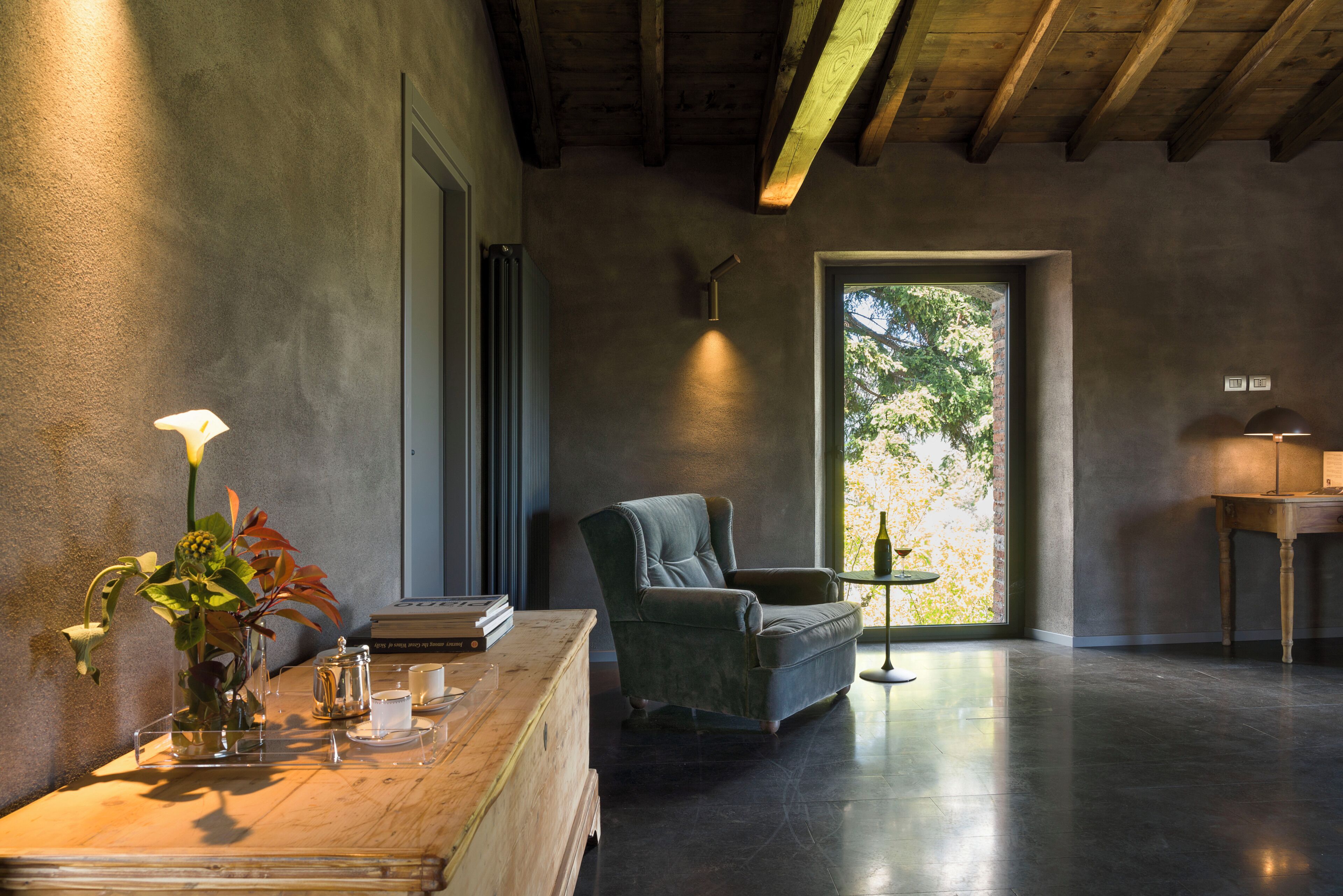 Una habitación con techo de madera, un sillón verde, una mesita auxiliar con un florero y vistas a un espacio exterior por una puerta.