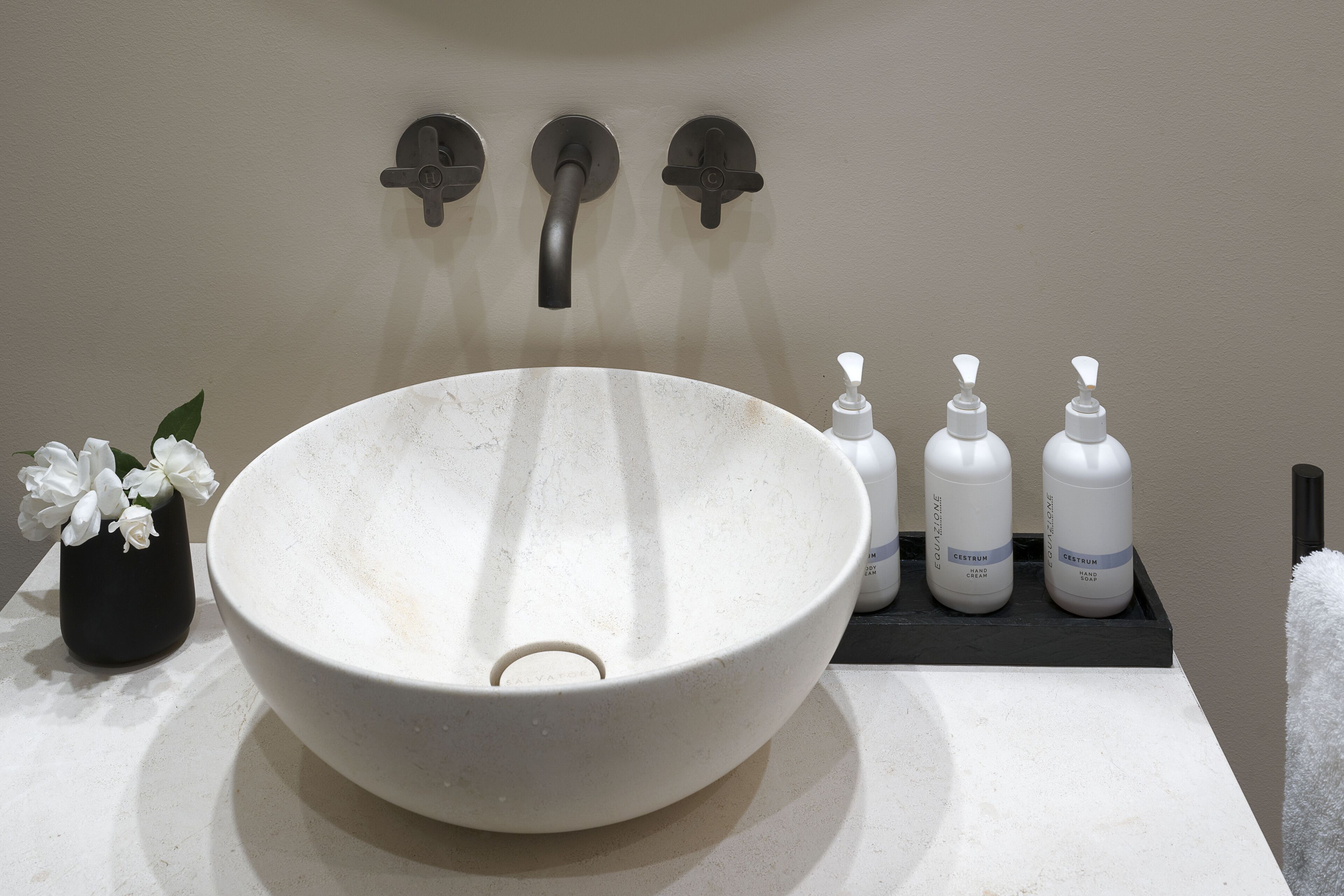 A bathroom with a large white vessel sink, a black vase with white flowers, and three bottles of hand cream on a black tray.