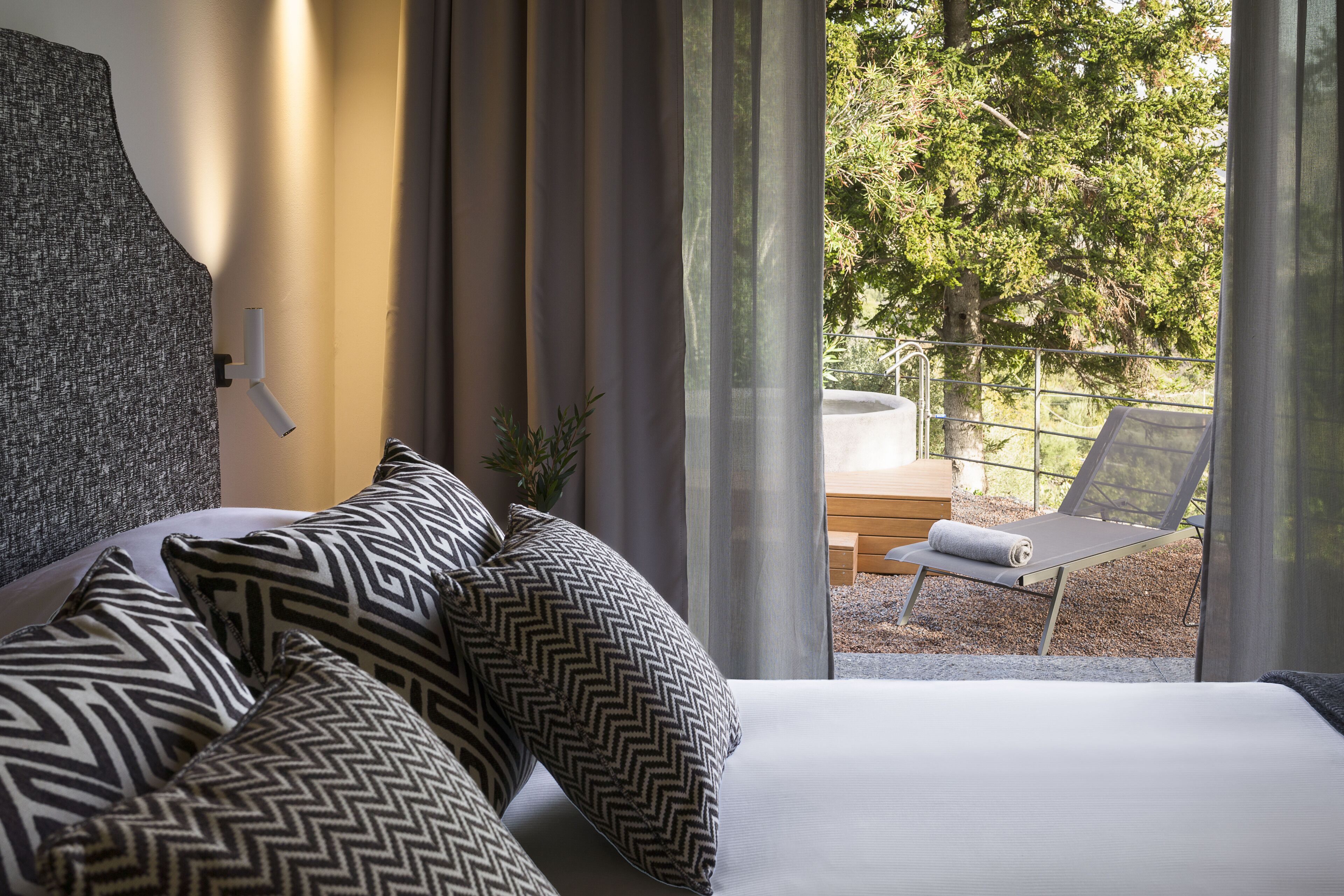 A bedroom with a bed, patterned pillows, a view of an outdoor seating area with a chair and table.