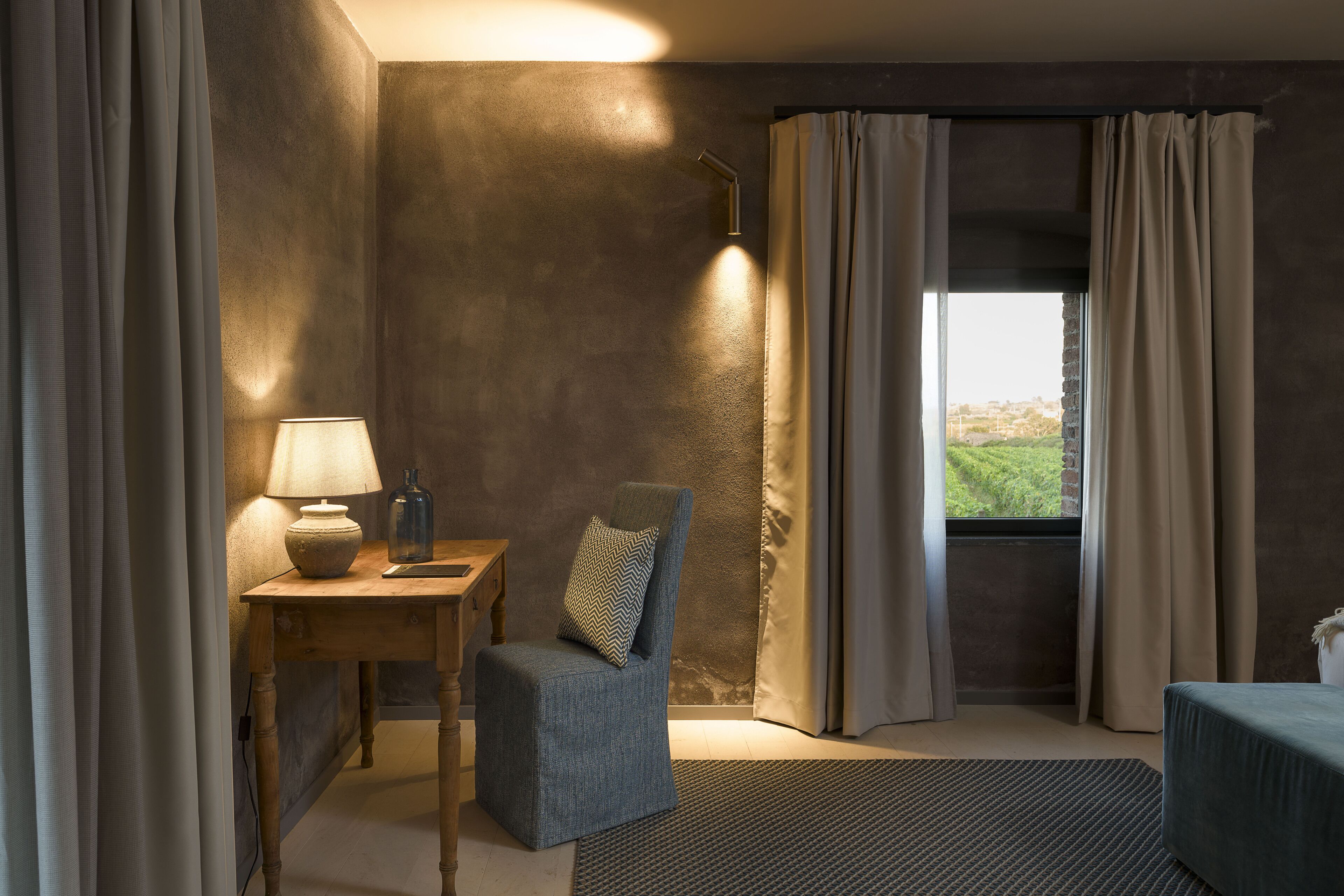 A room with a wooden desk, a chair, a lamp, and a view of greenery through a window.
