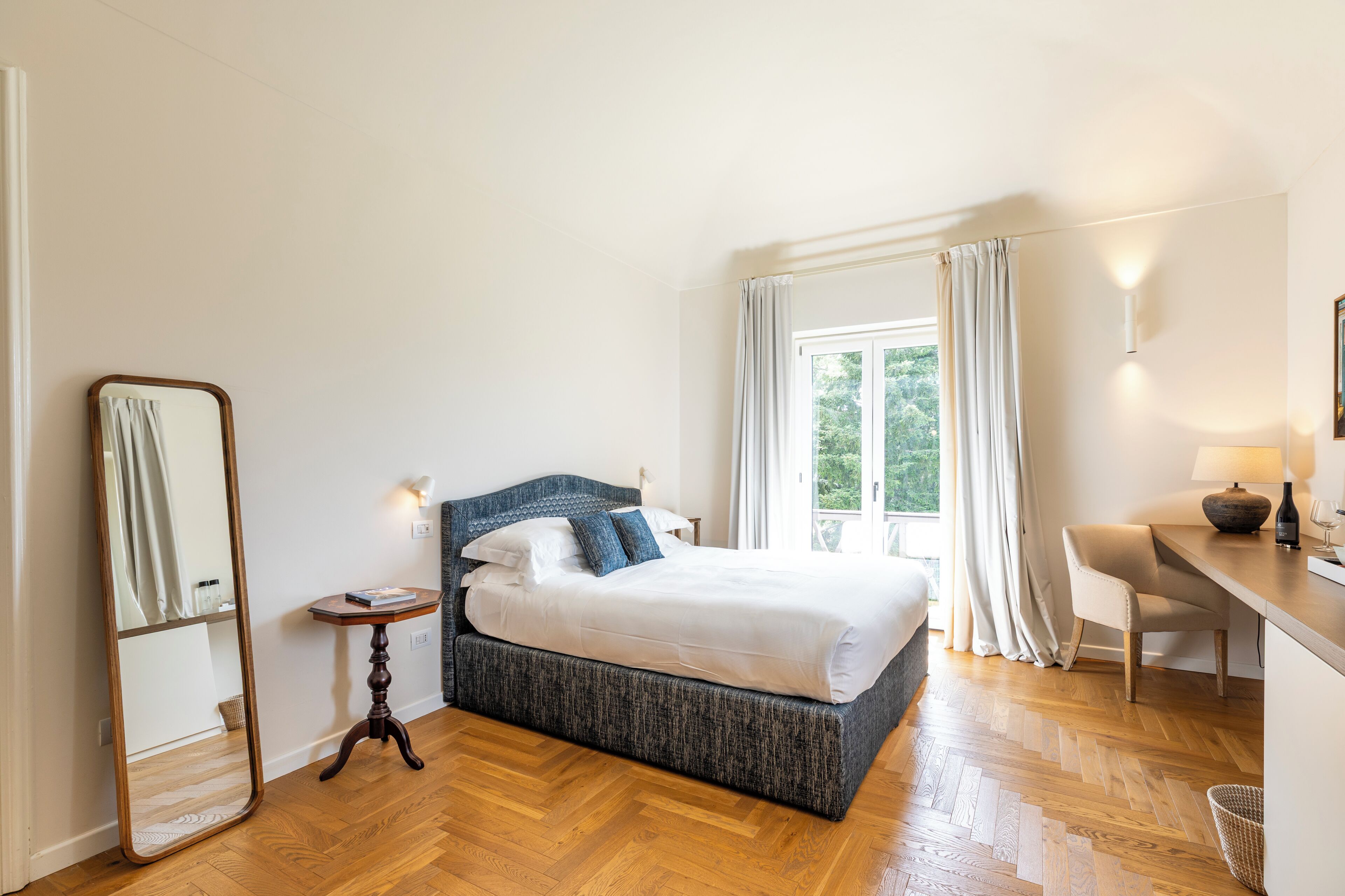 Une chambre à coucher comprenant un lit, une table de chevet, une chaise, un bureau, un miroir et une fenêtre avec des rideaux.