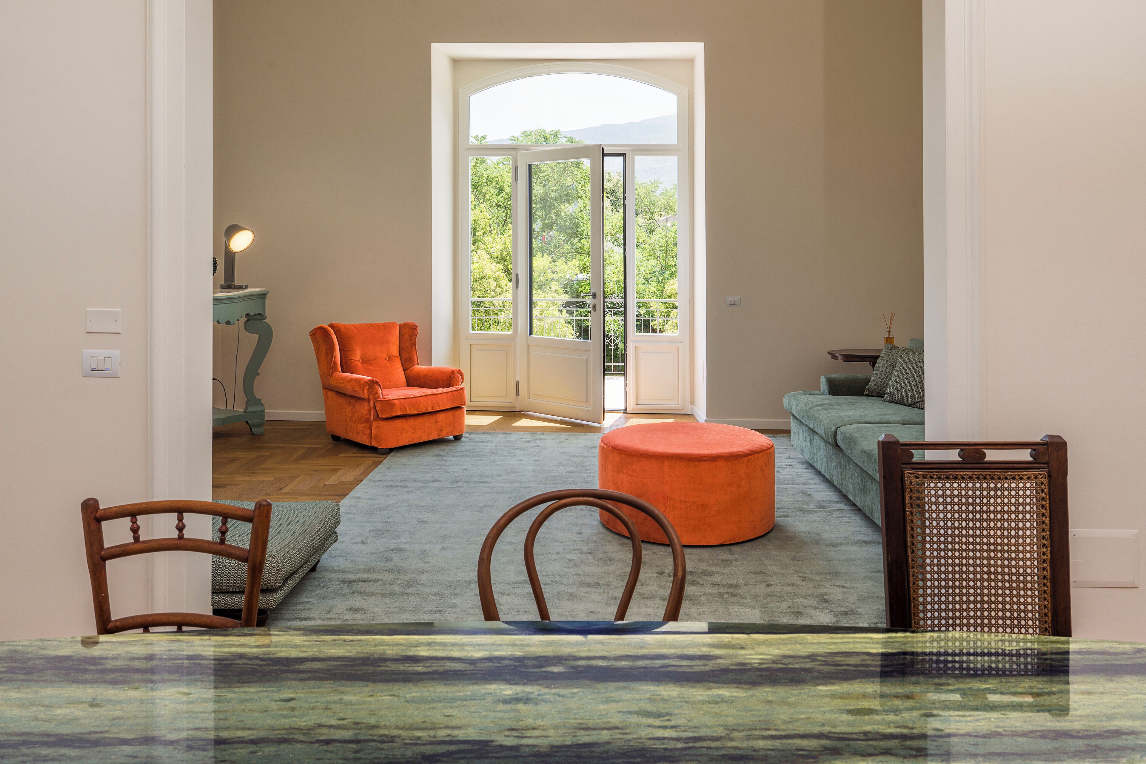 A spacious living room with a large window, a sofa, an orange armchair, and a wooden chair.