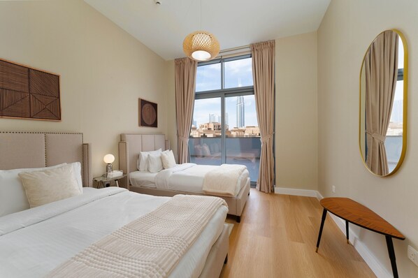 A hotel room with two beds, a large window with curtains, a round mirror, and a wooden bench.