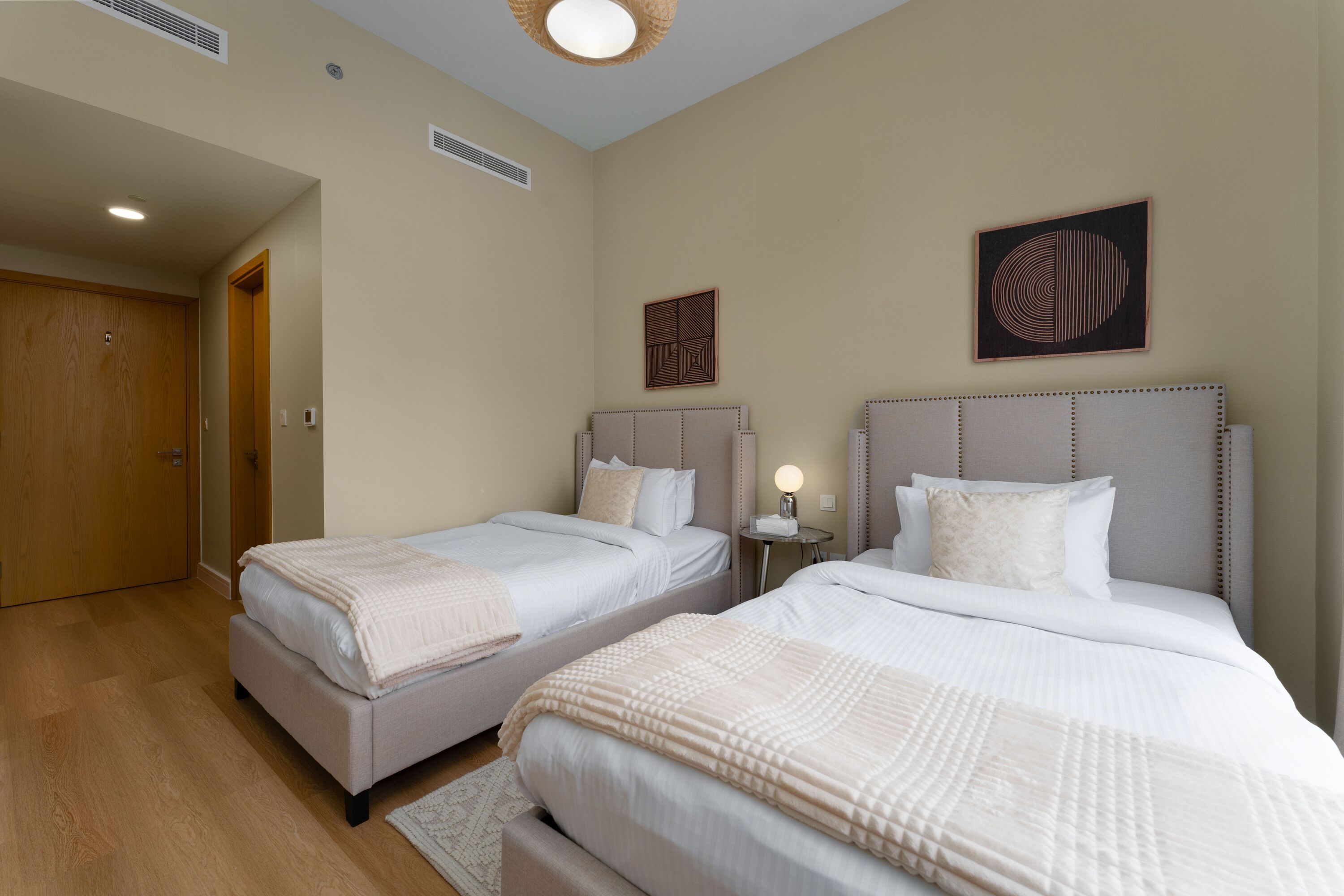 A hotel room with two beds, a wooden door, a small table with a lamp, and a wall-mounted artwork.