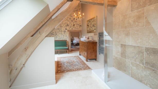 Een kamer met hoge plafonds en een schuine wand, een houten bank, een groene fauteuil, een houten dressoir, een spiegel en een douchehoek.