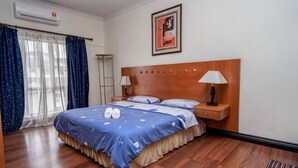 A hotel room with a wooden bed, a nightstand with a lamp, a wall-mounted air conditioner, and a framed picture above the bed. - Nana Marina Court (Kota Kinabalu)