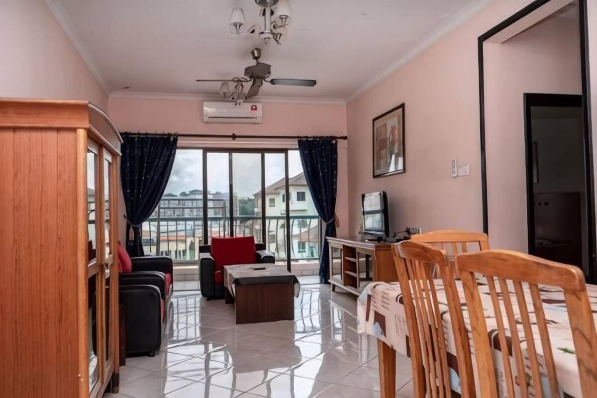 Een woonkamer met een bank, een eettafel, een televisie en een balkon met gordijnen.