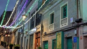 Una strada fiancheggiata da edifici, alcuni con balconi e persiane verdi, illuminata da una ghirlanda di luci multicolori.