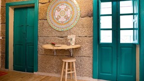 Una stanza con una parete in pietra, due porte color petrolio, un tavolo rotondo con un piatto e uno sgabello, e una finestra con vista.