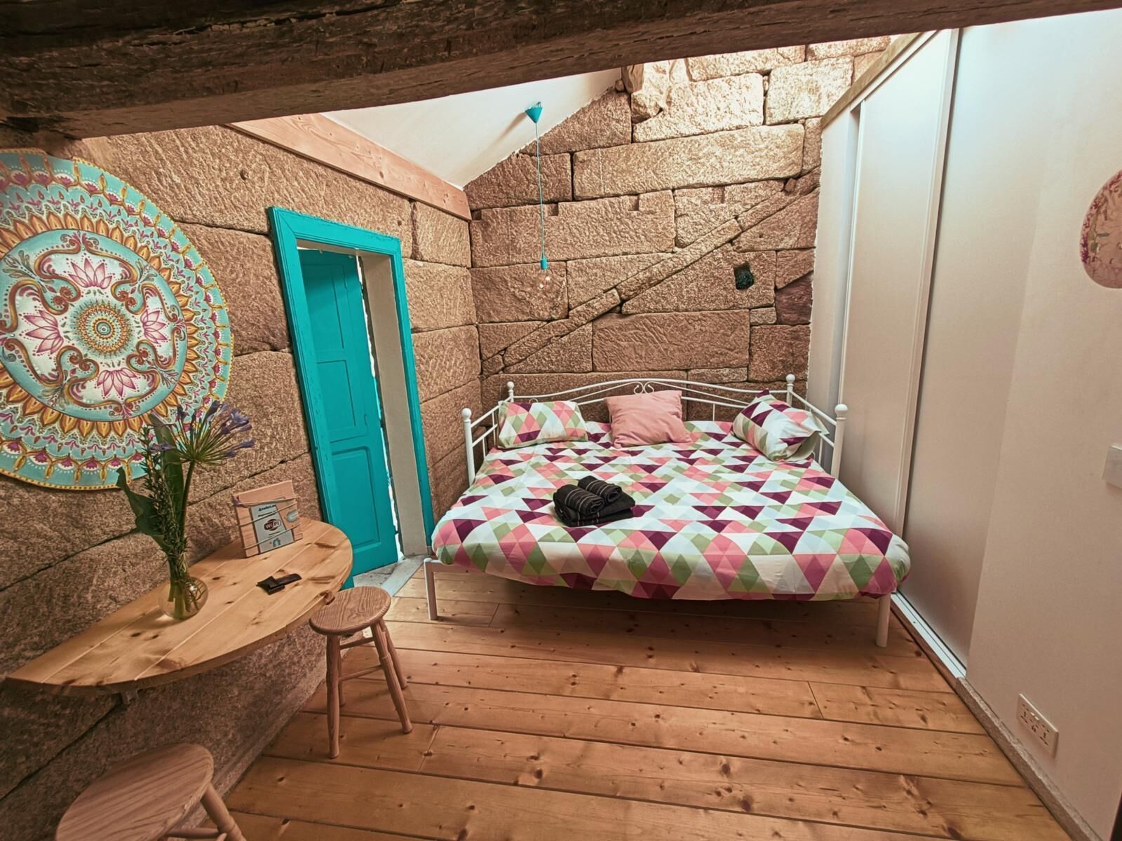 Un dormitorio acogedor con pared de piedra, puerta azul, cama con colcha de colores, mesa de madera y un taburete pequeño.