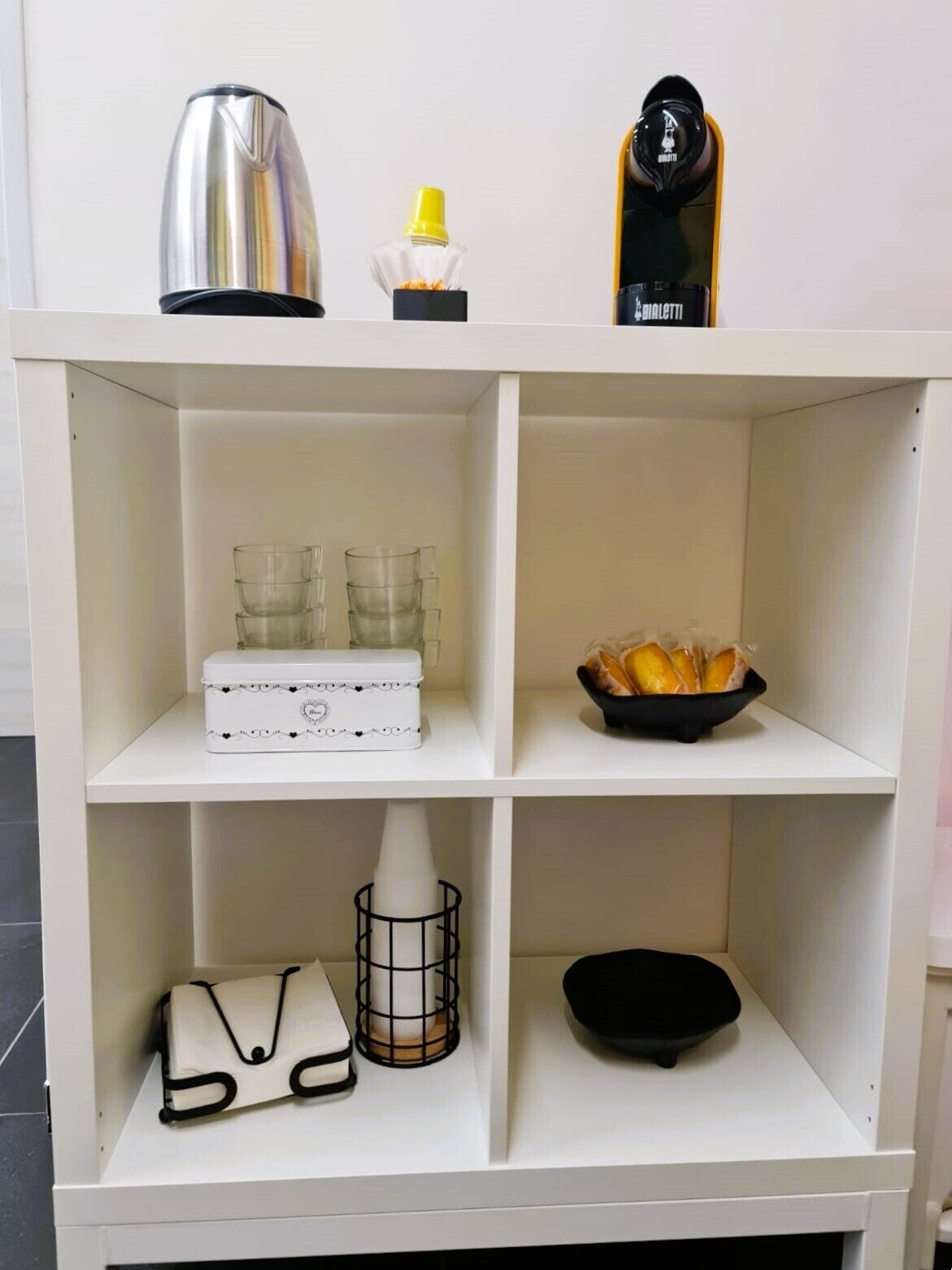 A white shelving unit with various items: a stainless steel kettle, a yellow bottle, a black object with a white label, a white box with a decorative design, a black bowl with bread, a white handbag, and a black bowl.