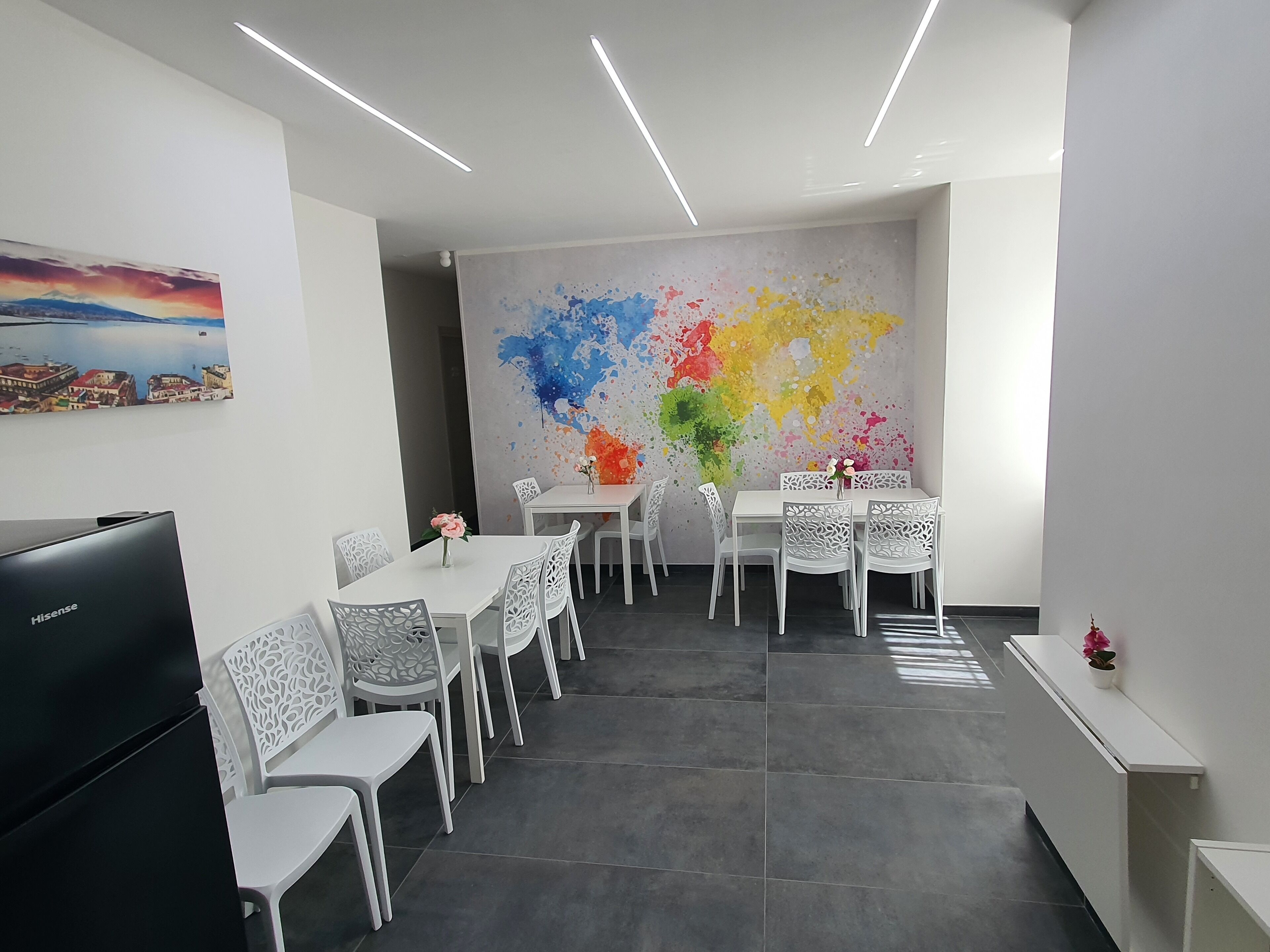 Een moderne eetruimte met witte tafels en stoelen, een groot abstract schilderij aan de muur, en een flatscreen televisie aan de linkerkant.