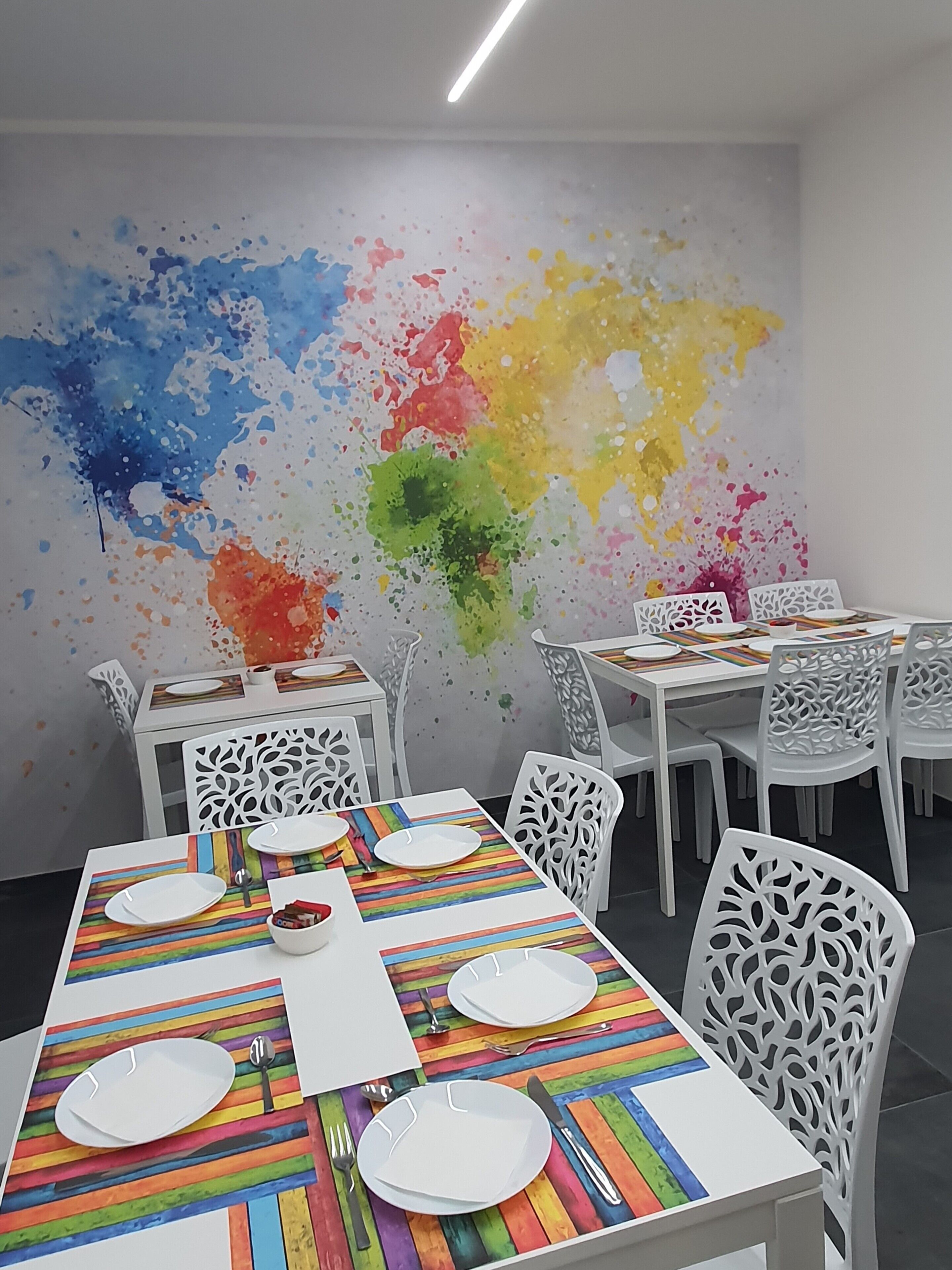 A dining area with white chairs and tables, each set with white plates and cutlery. A colorful abstract wall art is visible in the background.