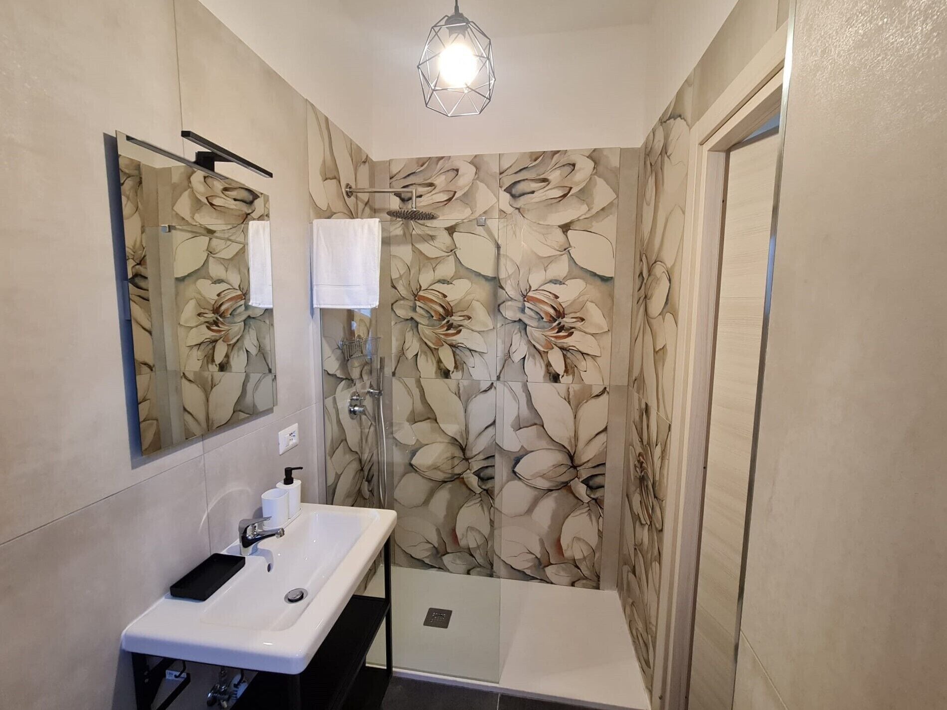 Une salle de bain avec un lavabo blanc, un miroir, une douche avec une porte en verre et un papier peint à motifs floraux.