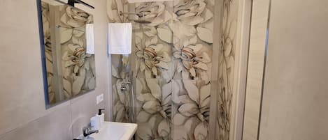 Une salle de bain avec un lavabo blanc, un miroir, une douche avec une porte en verre et un papier peint à motifs floraux.