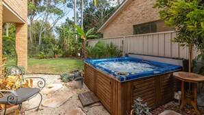 A hot tub in a backyard with a brick house, a wooden deck, and various plants. - 21 Cedar Grove - Pet Friendly (Forster)