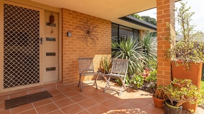 A covered patio with a brick wall, two wooden chairs, and potted plants. - 21 Cedar Grove - Pet Friendly (Forster)