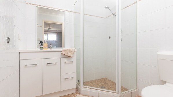 Een badkamer met een douchecabine van glas, witte betegelde wanden, een wit wastafelmeubel met spiegel en een toilet.