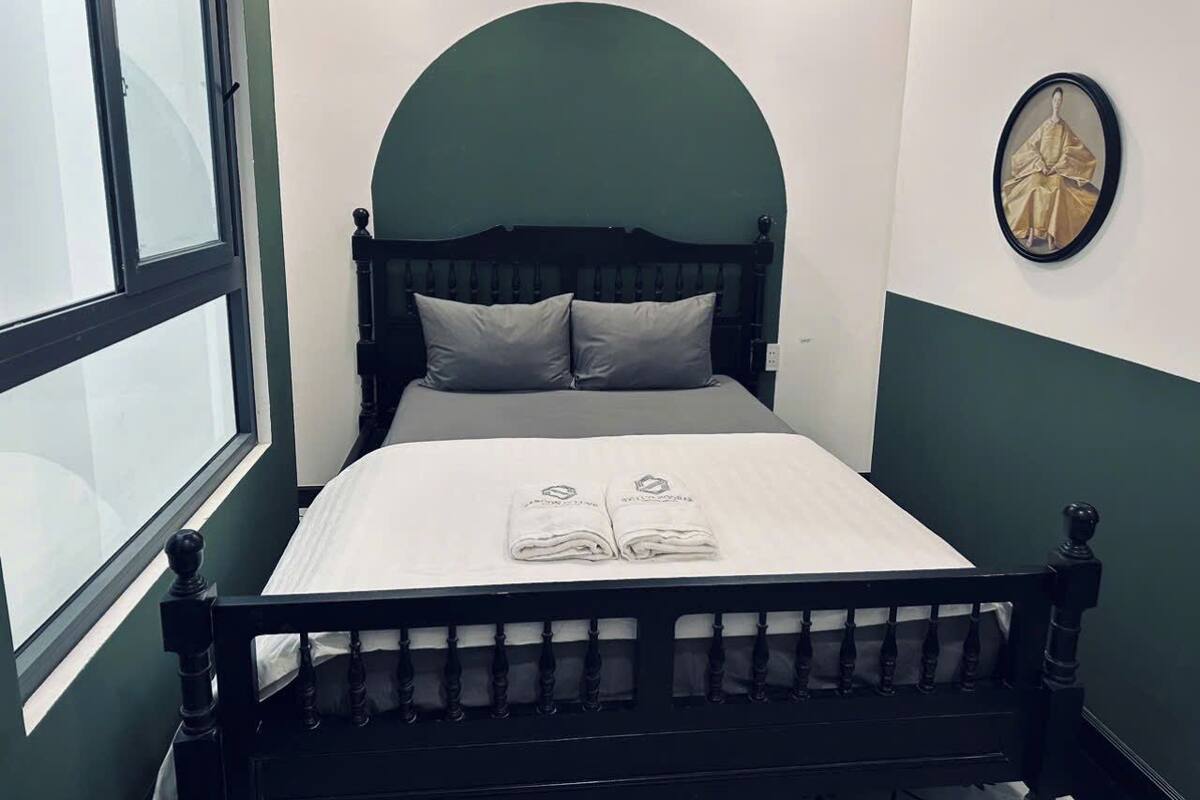 Uma cama de solteiro com estrutura de madeira escura, colchão branco e duas almofadas. Uma janela com persianas é visível à esquerda.