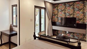 A modern room with a TV on a black stand, a wicker chair, a small table with a vase, and a window with curtains. - An Living (Ho Chi Minh City)