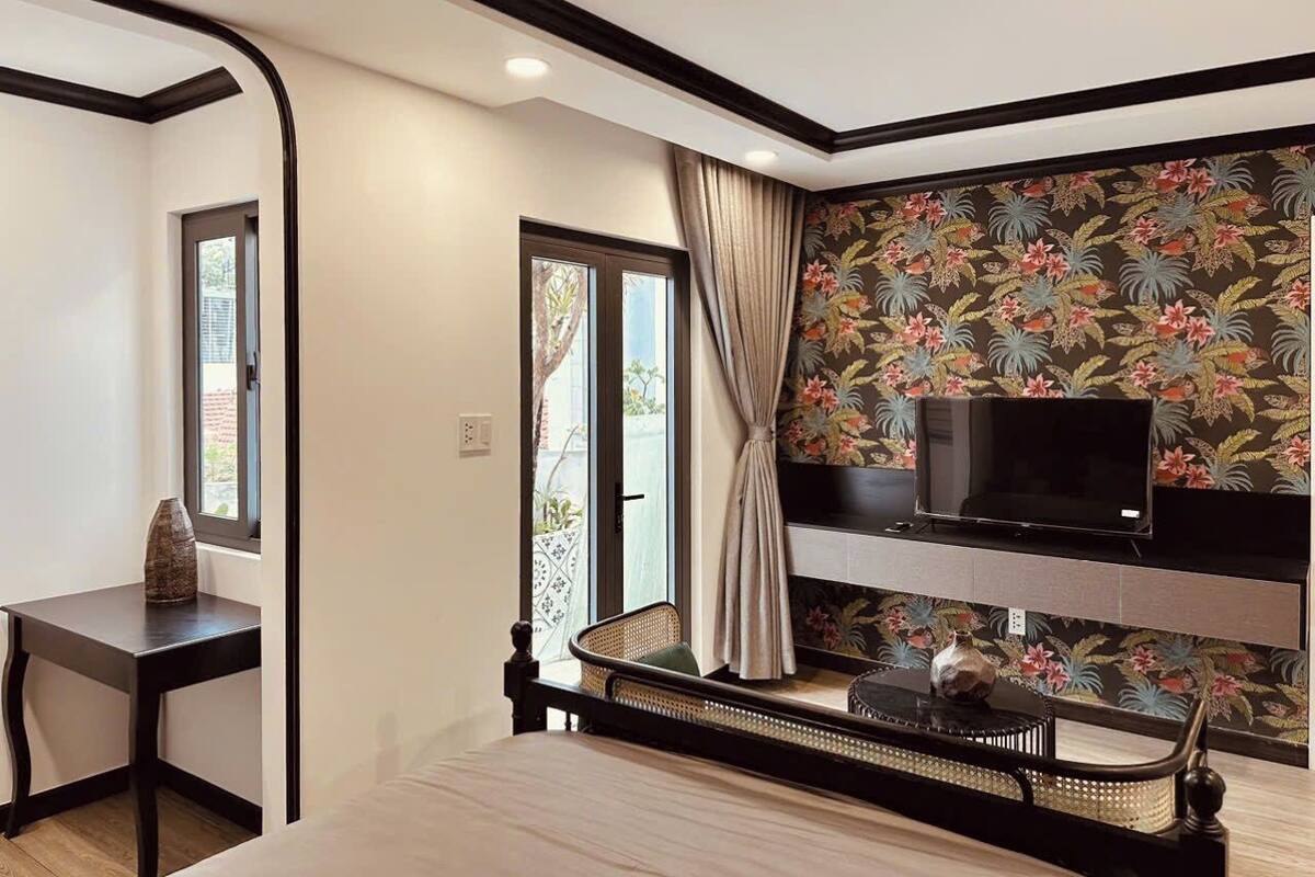 Quarto moderno com TV em suporte preto, uma cadeira de vime, uma pequena mesa com um vaso e uma janela com cortinas.