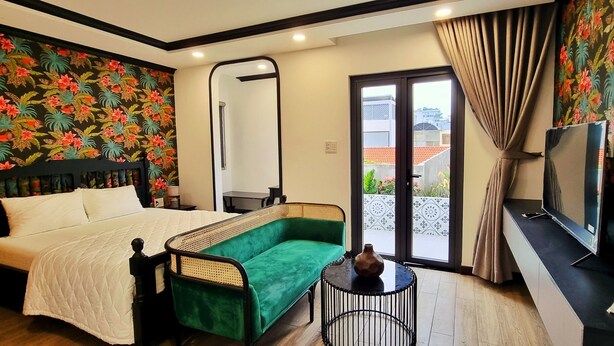A hotel room with a bed, a green sofa, a small table with a vase, a television, and a sliding glass door to the outside. - An Living (Ho Chi Minh City)