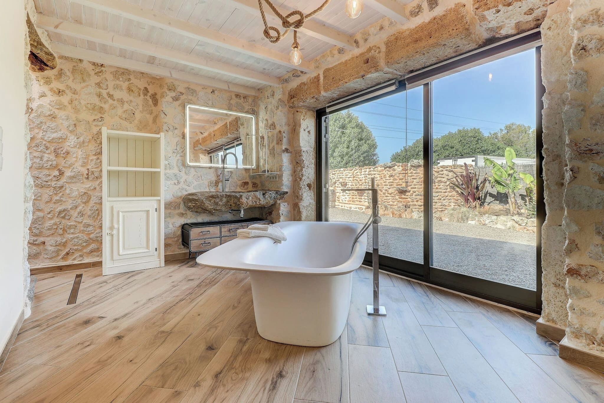 A bathroom with a freestanding bathtub, stone walls, and a large window offering a view of the outdoors.
