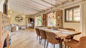 A spacious room with a wooden ceiling, a dining table with chairs, a fireplace, and a large window. - Petit Bijouc by Rentallorca (Can Picafort)