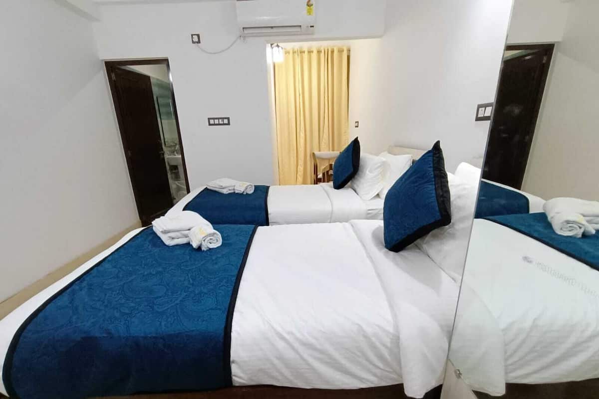 A hotel room with two beds, each with a blue bedspread and white linens, a small table with a chair, and a window with curtains.