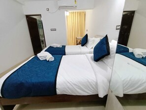 Quarto de hotel com duas camas, cada uma com colcha azul e roupa de cama branca, uma mesinha com uma cadeira e uma janela com cortinas.