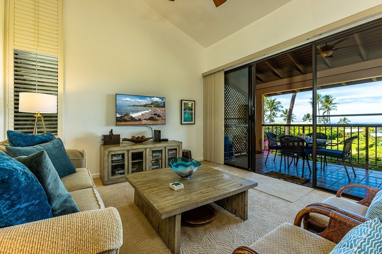 A living room with a sofa, coffee table, and a television. There is a balcony with outdoor furniture and a view of palm trees.