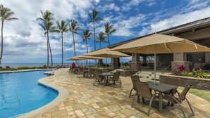 A poolside area with lounge chairs and umbrellas, a stone-tiled patio, and a view of the ocean with palm trees. - Wailea Ekahi One Bedrooms - Ocean View by Coldwell Banker Island Vacations (Kihei)