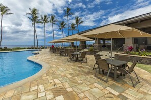 Ein Poolbereich mit Liegestühlen und Sonnenschirmen, eine terrasse mit Steinplatten und Blick auf das Meer mit Palmen.